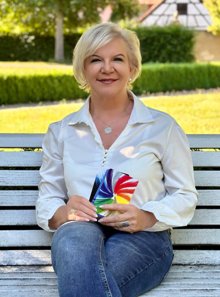 Petra Straetz sitzend auf Parkbank. Bild gerade von vorne. Sonniger Tag. Petra laechelt und haelt Karten in der Hand.